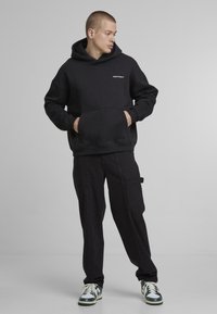 Black hoodie with a kangaroo pocket, made of soft fabric. Paired with black trousers and green-tinged sneakers, creating a monochromatic look.