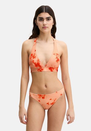 Donna che indossa un bikini halter color pesca con stampe floreali arancioni vivaci, in piedi davanti a uno sfondo semplice di colore grigio chiaro.