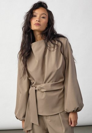 Femme aux longs cheveux foncés porte un haut beige cache-cœur à manches larges et un pantalon assorti, debout devant un fond clair uni.