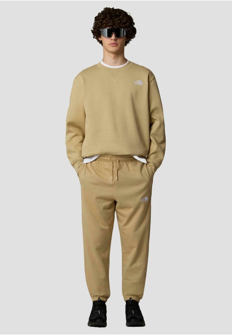 Sweatshirt beige et jogging assorti avec une taille élastique, présentant un petit logo blanc sur la poitrine gauche et la cuisse droite. Baskets noires.