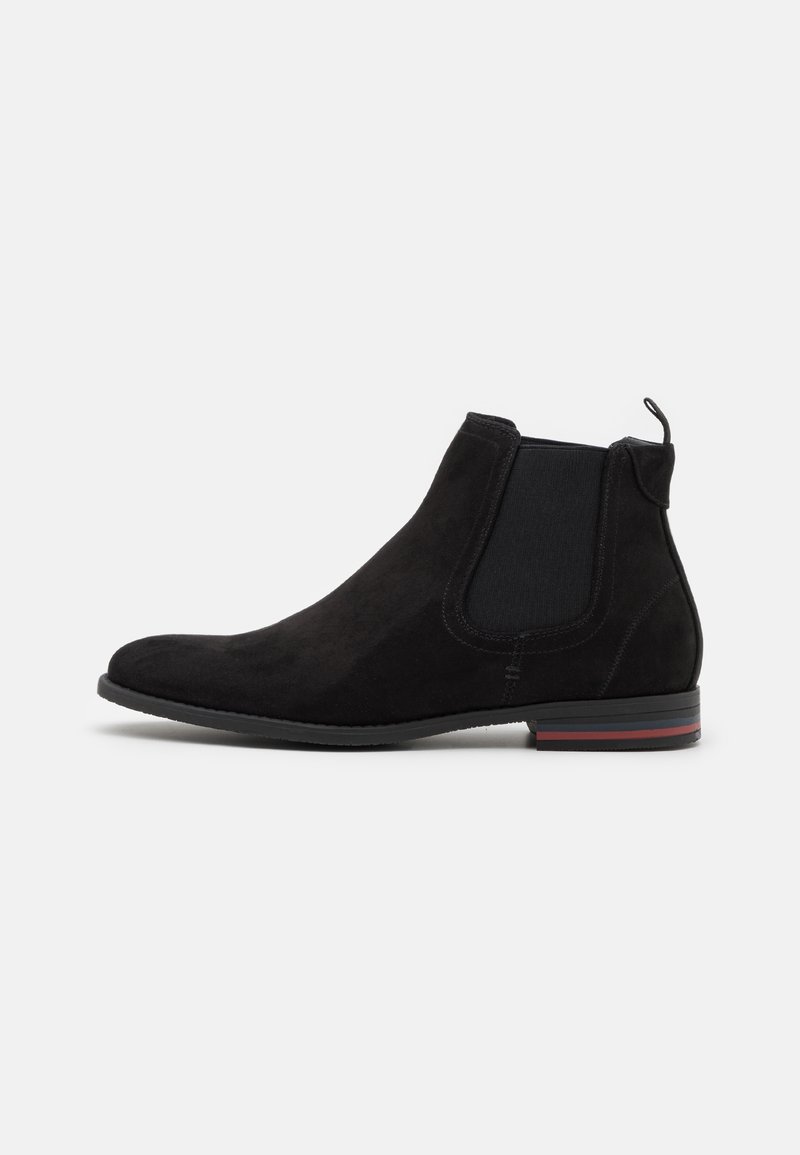 Botte Chelsea en daim noir avec panneau latéral élastique, languette à l'arrière et semelle plate ornée de bandes rouges et grises.
