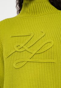 Zöld bordázott kötött garbó pulóver, amelyen mellrészen egy hozzáillő zöld, texturált madzag absztrakt mintát alkot.