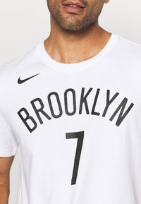 Valkoinen puuvillapaita, jossa on "BROOKLYN" lihavilla mustilla kirjaimilla ja numero "7" perforoidulla mustalla, pienen mustan Nike-logon korostamana.