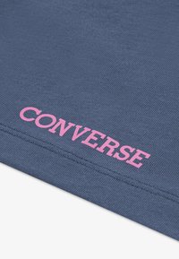 Tejido azul oscuro con una textura suave, que presenta el gran texto en rosa "CONVERSE" aplicado de manera destacada. Vista en primer plano del detalle del logo.