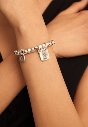 Pulsera de cuentas plateadas con dos dijes, uno grabado con "SUERTE", llevada en una muñeca contra un fondo negro.