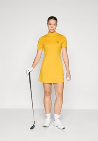 Žlté golfové šaty s vysokým golierom, krátkymi rukávmi a kruhovým logom. Modelka drží golfovú palicu, má na sebe biele topánky a rukavice. Jednoduchý dizajn.