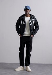 Black lightweight jacket with "ICON" print, white sweater, black slim-fit pants, and beige sneakers. Blue denim cap completes the outfit.