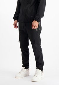 Zwarte cargobroek met zijzakken, elastische tailleband en trekkoord. Gepaarde met witte sneakers, voorzien van een gladde stofstructuur.