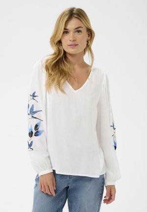 Žena s dlhými blond vlasmi má na sebe bielu blúzku s výstrihom do V, zdobenú modrou výšivkou vtáčika a listov na rukávoch, a svetlomodré džínsy.