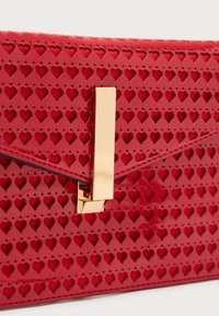Pochette en cuir verni rouge avec découpes en forme de cœur, dotée d'une fermeture à charnière dorée et d'un design élégant en forme d'enveloppe. Surface texturée.