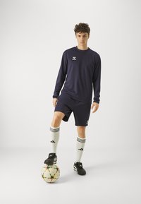 Camicia a maniche lunghe blu scuro e pantaloni corti con logo bianco, abbinata a calze bianche con tre strisce nere, in piedi su un pallone da calcio multicolore.