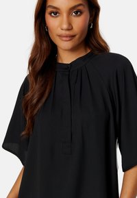 Blusa nera con colletto alla cinese, chiusura a bottoni e design plissettato. Presenta maniche ampie e una texture di tessuto liscia per il massimo comfort.