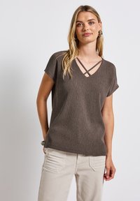 Haut court à manches courtes texturé marron avec un col en V et des bretelles croisées. Associé à un pantalon beige clair, présentant une coupe décontractée.