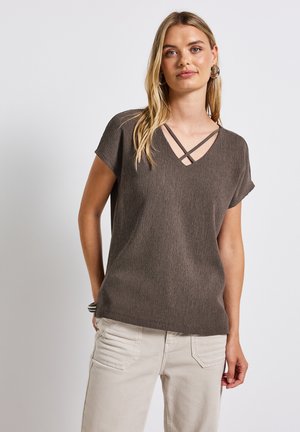 Brun texturerad kortärmad topp med V-ringning som har korslagda remmar. Matcher med ljusbeige byxor, som visar en avslappnad passform.