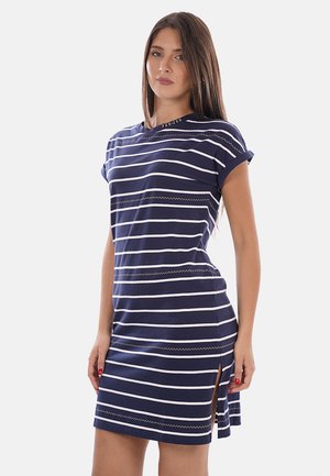 Donna che indossa un vestito a maniche corte con righe orizzontali blu navy e bianche con spacco laterale, in piedi davanti a uno sfondo bianco semplice.