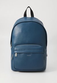 Blå læderrygsæk med en rund form, lomme med lynlås foran og topgreb. Har diskret branding på frontpanelet.