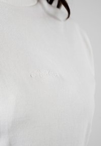 Biały prążkowany golf z delikatnie wytłoczonym logo. Materiał ma miękką fakturę i dopasowany krój.