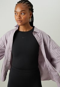 Light purple striped oversized shirt made of cotton, worn over a black fitted bodysuit with a subtle Nike logo. Earrings are round and transparent.
