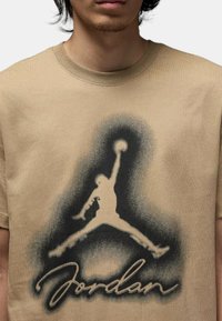 T-shirt in cotone beige con una silhouette nera di un atleta che salta e il testo "Jordan", arricchita da un effetto spray a gradiente attorno al design.