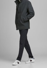 Cappotto di lana grigio con colletto alto, tasca sul petto singola e chiusura con bottoni, abbinato a pantaloni blu navy e sneakers bianche con dettagli rossi e blu.