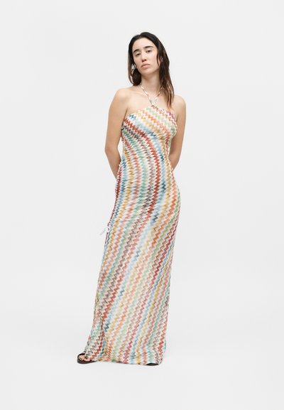 Γυναίκα που φοράει μια maxi αμάνικη φόρεμα με πολύχρωμο σχέδιο ζιγκ-ζαγκ, στέκεται με τα χέρια πίσω από την πλάτη της μπροστά σε απλό φόντο.