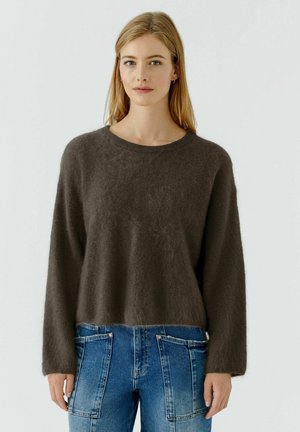 Blonde woman wearing a loose dark brown fuzzy sweater and blue denim jeans with large front pockets, standing against a plain background.