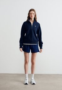 Γυναίκα που στέκεται φορώντας μπλε σκούρο φούτερ με φερμουάρ στο μισό μήκος, ταιριαστά σορτς, λευκές κάλτσες και λευκά αθλητικά παπούτσια, σε απλό φόντο.