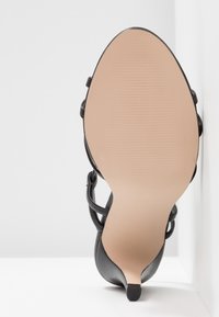 Sandale noire à talons hauts avec un dessus lisse, bout ouvert et fines lanières. Semelle beige texturée avec un léger rainurage, talon pointu.