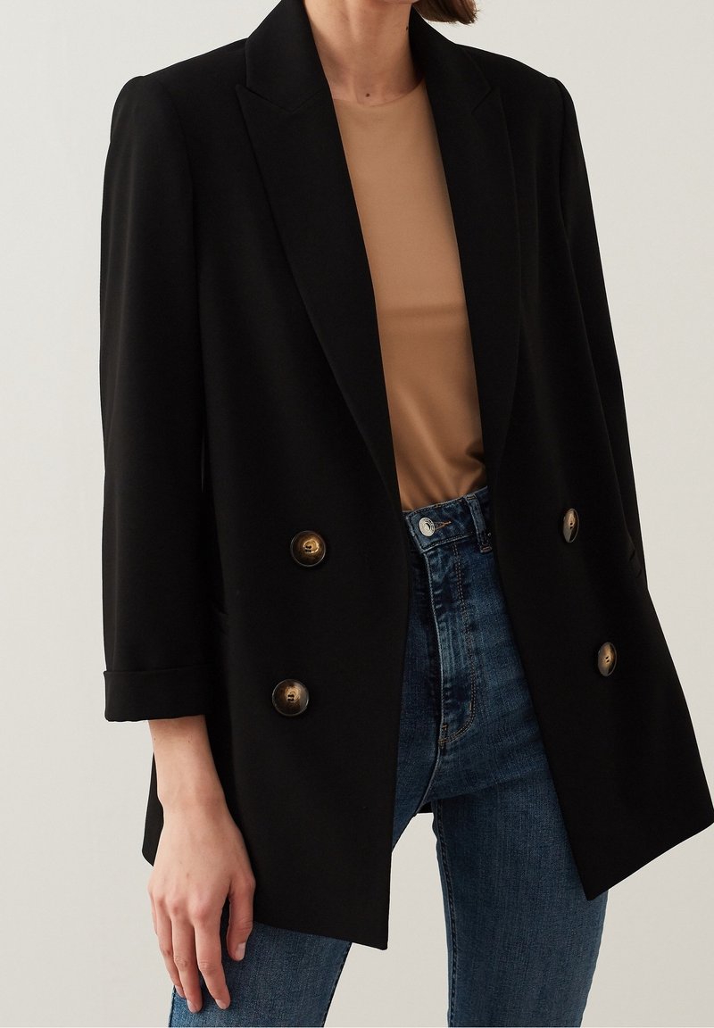 Blazer negro de corte cruzado con botones dorados, diseño ajustado, hombreras y puños doblados, combinado con una blusa beige y jeans azules.