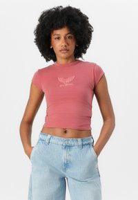 Kortermet, tettsittende, støvete rosa T-skjorte med en logo på forsiden. Brukt sammen med høyhalsede, lyseblå denimjeans.