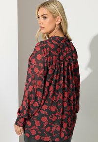 Sort gennemsigtig bluse med rødt blomstermotiv, lange ærmer og samlet bagdetalje. Glat tekstur og løst fit.
