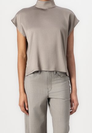 Haut sans manches taupe à col roulé, associé à un pantalon droit taille haute gris clair sur une personne debout devant un fond blanc.