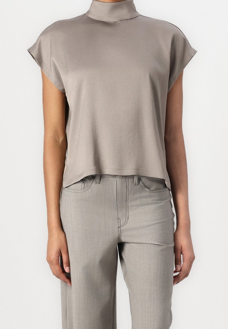 Haut sans manches taupe à col roulé, associé à un pantalon droit taille haute gris clair sur une personne debout devant un fond blanc.