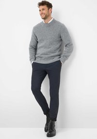 Uomo che sorride, indossando un maglione grigio a maglia, una camicia bianca, pantaloni scuri slim-fit e stivali neri con lacci, appoggiato a un muro bianco.