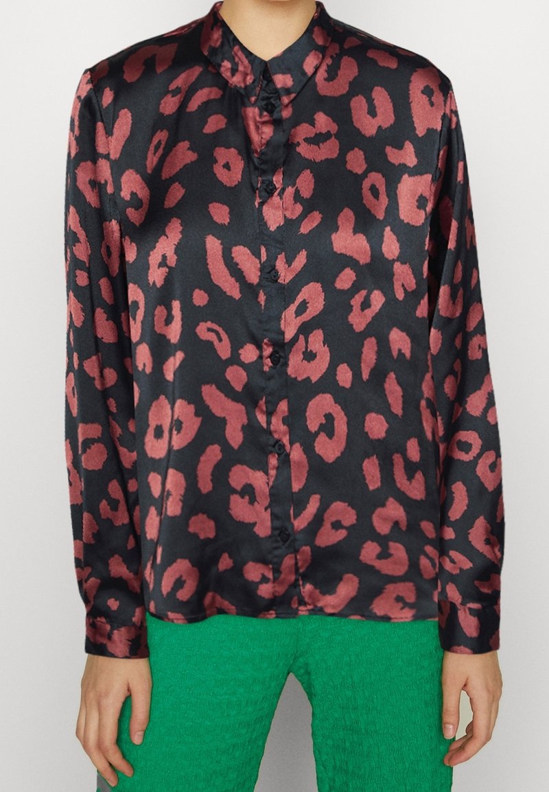 Blouse noire en satin à manches longues avec taches léopard rouge foncé, devant boutonné, associée à un pantalon vert vif texturé.