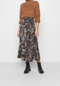 Un pull en tricot marron à manches 3/4 associé à une jupe midi noire avec un motif paisley coloré, porté avec des bottes noires à lacets.