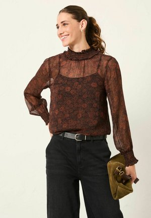 Durchsichtige schwarze Bluse mit floralem Muster, Rüschensaum am hohen Kragen und langen Ärmeln, kombiniert mit hoch taillierten schwarzen Jeans und einer grünen Wildleder-Clutch.