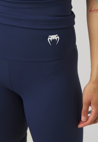 Marineblauwe sportlegging van gladde stof, met een hoge tailleband en een wit logo van een slang op de heup. Eenvoudig ontwerp, zonder patronen.