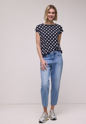 Ung kvinde står, iført en sort og hvid prikket top, lyseblå jeans og sølvfarvede sneakers, smilende mod en ensfarvet baggrund.