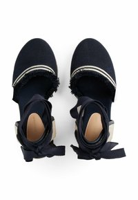 Navy blue espadrille shoes with a textured upper, frayed fabric accents, flat sole, and ankle ties. Features contrasting white stripes.