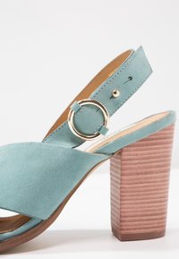 Sandale en daim bleu sarcelle avec une large bride et une boucle circulaire dorée. Elle présente un talon carré à rayures multiples roses offrant une texture et une stabilité supplémentaires.