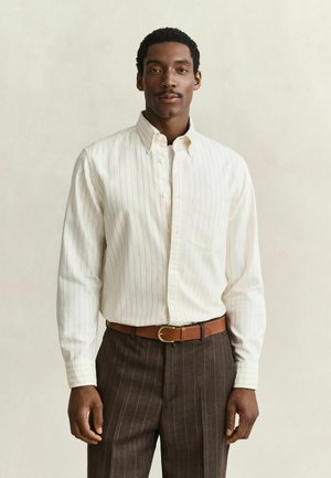 Man draagt een wit gestreept overhemd met knopen, in een bruine gestreepte broek met een bruine riem, staand tegen een effen achtergrond.