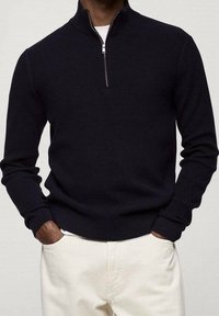 Dunkelblaues geripptes Pullover mit einem Viertel-Reißverschluss-Kragen, langärmlig und mit einem engen Saum. Getragen über cremefarbenen Hosen.