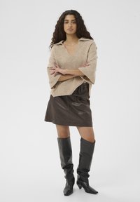 Pull beige en maille avec un col en V et des manches larges, associé à une mini-jupe en cuir marron foncé et des bottes noires hautes jusqu'au genou avec des bouts pointus.