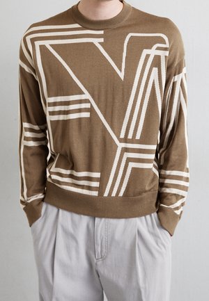 Homme portant un pull marron avec de grandes lettres géométriques blanches et des motifs, assorti à un pantalon gris clair, mains dans les poches.