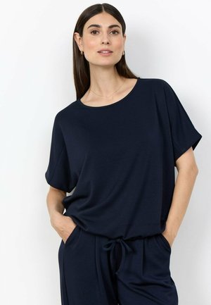 Vrouw met lang donker haar, gekleed in een marineblauw kortemouwen top en bijpassende broek, met handen in de zakken tegen een witte achtergrond.