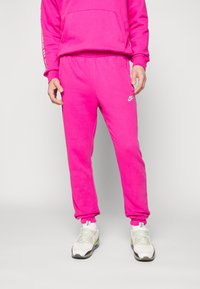 Osoba v jasně růžových Nike teplácích a odpovídající mikině, doplněná bílými a světle zelenými Nike teniskami, stojící na neutrálním pozadí.