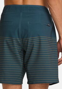 Herren-Badehosen in Petrol mit horizontalen senfgelben Streifen. Verfügt über eine Gesäßtasche und ein Etikett am Bund. Glatte Textur, mittlere Leibhöhe.