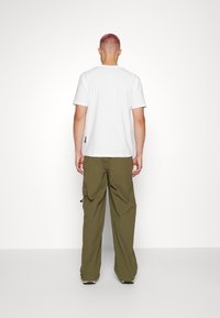 Person with short pink hair stands facing a white wall wearing a plain white t-shirt, olive green cargo pants, and light-colored shoes.