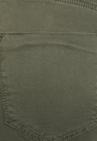 Olive green denim fabric featuring a back pocket with contrasting stitching. Smooth texture with a slight sheen, no visible patterns or embellishments.